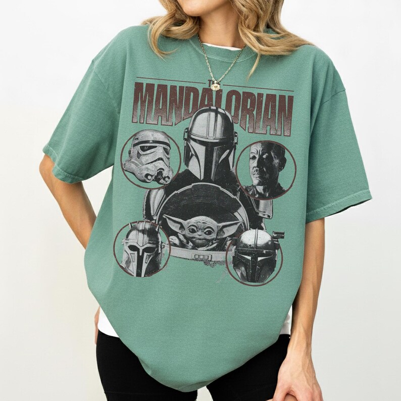 Pu&ograve; includere: T-shirt verde con un design grafico con personaggi di The Mandalorian. Il design include il testo "MANDALORIAN" e immagini di un casco da Stormtrooper, The Mandalorian, Grogu e altri personaggi.