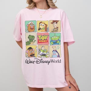 May include: A light pink t-shirt featuring a grid of Toy Story characters, including Mr. Potato Head, Woody, and Buzz Lightyear. The shirt also has the text "Toy Story" and "Walt Disney World" printed on it.