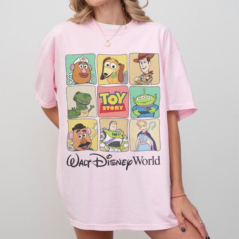 May include: A light pink t-shirt featuring a grid of Toy Story characters, including Mr. Potato Head, Woody, and Buzz Lightyear. The shirt also has the text "Toy Story" and "Walt Disney World" printed on it.