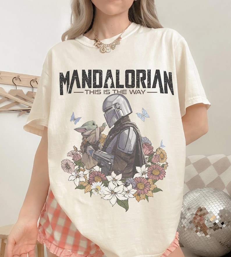 Op de afbeelding: Cr&egrave;mekleurig T-shirt met de zwarte tekst "MANDALORIAN - THIS IS THE WAY". De afbeelding toont de Mandalorian en Grogu omringd door bloemen en vlinders. Het shirt heeft korte mouwen.