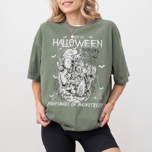 Puede incluir: Camiseta verde oliva con un dise&ntilde;o de Halloween. El dise&ntilde;o presenta una colecci&oacute;n de personajes espeluznantes, murci&eacute;lagos y telara&ntilde;as, con el texto "This Is Halloween" y "Nightmare on Mainstreet" en una fuente desgastada.