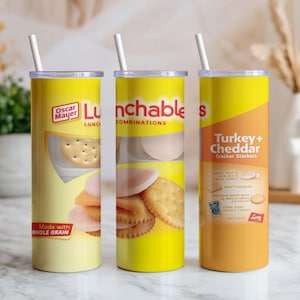May include: Three cylindrical tumblers featuring a yellow and orange design inspired by Lunchables Turkey and Cheddar Cracker Stackers packaging. Each tumbler has a clear lid, a white straw, and displays graphics of crackers, meat, and cheese.