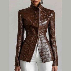 May include: A dark brown jacket with a glossy crocodile skin texture, featuring a high collar, long sleeves, and a single front button closure. The jacket is fitted at the waist and is paired with plain white trousers, creating a structured look.