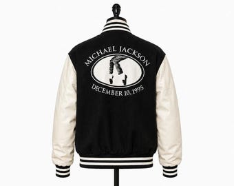 May include: A black varsity jacket with cream-colored sleeves, featuring a ribbed collar, cuffs, and hem with white stripes. The back displays a white oval graphic with the text MICHAEL JACKSON and DECEMBER 10, 1995, surrounding a silhouette of legs.