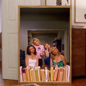 Op de afbeelding: Een grote, rechthoekige spiegel met een houten frame, die een kamer met vier vrouwen weerspiegelt. De spiegel staat op een plank vol boeken. Twee ingelijste prints met luipaardprintletters hangen aan de muur. De kamer heeft hardhouten vloeren.