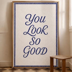 May include: A framed artwork featuring the phrase "You Look So Good" in blue script on a white mosaic background. The frame is gold, and the artwork is bordered with a blue mosaic trim. A small wooden side table is in the foreground.