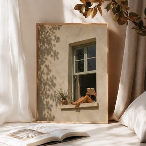 May include: A rectangular framed wall art print featuring a beige stucco wall with a sage green window frame. A person sits on the window ledge reading an open book. A small potted plant sits on the ledge. The scene is bathed in soft, warm sunlight.