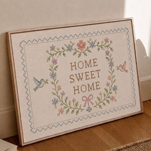Könnte beinhalten: Gerahmtes Kreuzstich-Kunstwerk mit dem Schriftzug "HOME SWEET HOME". Das Kunstwerk zeigt einen Blumenkranz mit rosa, blauen und grünen Blumen und zwei Vögeln. Der Rahmen ist hellbraun.