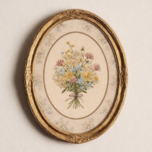 May include: An oval, gold-framed artwork featuring a floral embroidery. The embroidery depicts a bouquet of colourful flowers, including pink, yellow, and blue blooms, tied with a pink ribbon. The frame has ornate detailing.