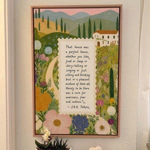 Op de afbeelding: Een ingelijste kunstdruk met een citaat van J.R.R. Tolkien, tegen een levendig landschap. De afbeelding toont een pad, een huis en heuvels met bomen en bloemen. Het citaat staat in een decoratief kader.