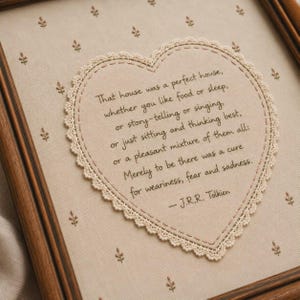 Op de afbeelding: Een ingelijste stoffen wandhanger met een hartvormig middenstuk en crèmekleurige kanten rand. De beige stof heeft een klein bloemenpatroon. De tekst luidt: That house was a perfect house, whether you like food or sleep, or story-telling or singing, or just sitting and thinking best, or a pleasant mixture of them all: Merely to be there was a cure for weariness, fear and sadness. - J.R.R. Tolkien.