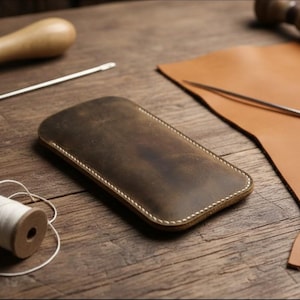 May include: A handcrafted, distressed brown leather phone case with white stitching, set on a wooden surface. The image also shows leather, needles, and thread, suggesting a leathercraft theme.