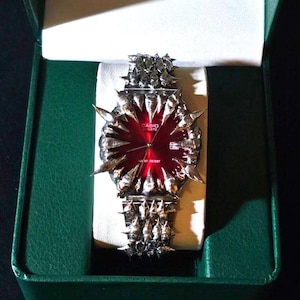 May include: A Casio Quartz watch with a red dial and silver-toned, spiked accents. The watch is displayed in a green box with a white interior. The watch band and bezel feature a spiky design.