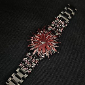 May include: A unique black watch with a striking red and black spiky design. The watch features a dark metal band and a central decorative element with a starburst appearance. The overall aesthetic is bold and edgy.