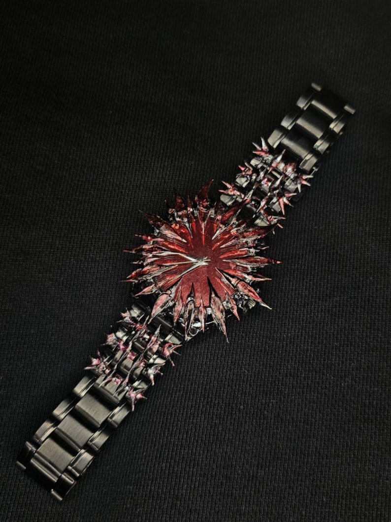 May include: A unique black watch with a striking red and black spiky design. The watch features a dark metal band and a central decorative element with a starburst appearance. The overall aesthetic is bold and edgy.