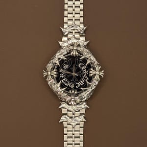 May include: Silver wristwatch with a black face and ornate cross and floral patterns. The watch has a stainless steel strap and a detailed bezel. A luxury item.