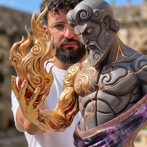 May include: A bust of a bearded figure with dark gray skin and gold accents, featuring a muscular arm extending to hold a sculpted gold flame. The base of the bust displays a purple and black nebula-like pattern with a gold Greek key border.