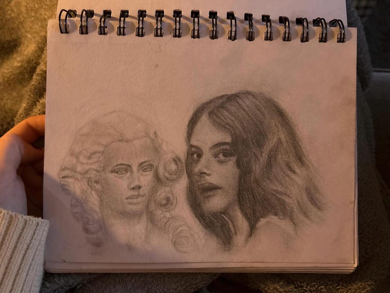May include: Pencil sketch of two female faces in a spiral-bound sketchbook. One face is a historical figure with a powdered wig, and the other is a modern woman with long, wavy hair. The drawing is in shades of grey.