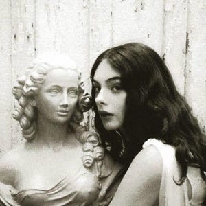 May include: Monochrome image showing a classical bust alongside a woman with dark, flowing hair. The woman is positioned close to the bust, creating a contrast. The background is a distressed, weathered wall, adding to the vintage aesthetic.