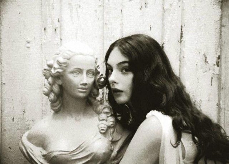 May include: Monochrome image showing a classical bust alongside a woman with dark, flowing hair. The woman is positioned close to the bust, creating a contrast. The background is a distressed, weathered wall, adding to the vintage aesthetic.