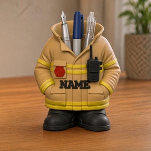 Könnte beinhalten: Ein beigefarbener Schreibtisch-Organizer in Form einer Feuerwehrjacke mit gelben Reflexstreifen, einem roten Emblem und einem schwarzen Funkgerät auf der Vorderseite. Das Wort NAME ist in schwarzen Buchstaben auf der Brust gedruckt. Er hält drei Stifte.