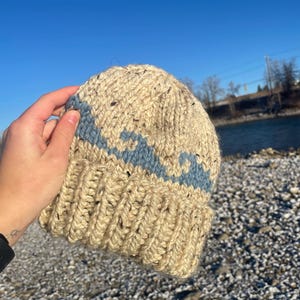 May include: A hand-knitted beanie in beige with flecks of dark brown. The hat features a light blue wave pattern across the crown and a ribbed brim. The hat is made of a thick, textured yarn, perfect for cold weather.