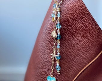 Könnte beinhalten: Ein goldfarbener Taschenanhänger mit Karabinerverschluss, der einen Muschelanhänger, einen Seestern und einen Herzanhänger aufweist. Der Anhänger enthält blaue und klare Perlen sowie weiße Perlen. Der Anhänger ist an einer braunen Ledertasche befestigt.
