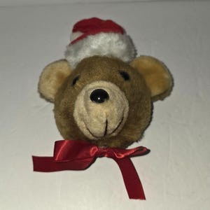 Puede incluir: Cabeza de oso de peluche marrón con gorro de Papá Noel rojo y blanco y lazo de cinta roja. El oso tiene nariz negra y orejas color canela. Decoración navideña festiva.
