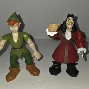 May include: Two plastic action figures of Peter Pan and Captain Hook. Peter Pan is in a green outfit with a brown belt and boots. Captain Hook is in a red coat with black boots and a hook hand. Both figures are standing.