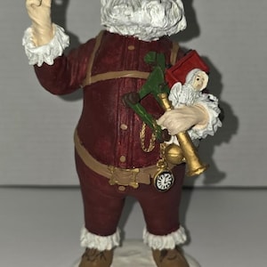 May include: A Santa Claus figurine wearing a deep red suit with tan straps and white fur trim. The figure holds a green toy, a small doll, and a gold pocket watch. The Santa has a white beard, a hat with holly, and brown boots, standing on a white base.
