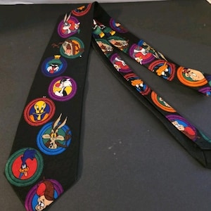 Puede incluir: Corbata negra con medallones de personajes de dibujos animados coloridos. Los medallones incluyen a Bugs Bunny, Pato Lucas, Piolín y otros personajes de Looney Tunes. La corbata es de tela lisa.