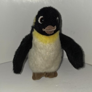 Könnte beinhalten: Ein Plüschpinguin-Spielzeug mit schwarzem Kopf und Flügeln, gelbem Hals und weißem Körper. Der Pinguin hat braune Füße und einen braunen Schnabel. Das Spielzeug steht aufrecht.