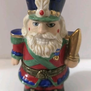 Puede incluir: Figura de cascanueces de cerámica con uniforme tradicional. La figura tiene barba blanca, ojos azules y uniforme rojo, verde y azul con detalles dorados. Sostiene un objeto dorado.