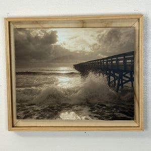 以下が含まれることがあります： セピア色の額縁に入った海の風景画。木製の桟橋が海に伸びています。波が雲の下で桟橋の支柱に打ち寄せ、太陽の光が雲を突き抜けています。