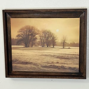 以下が含まれることがあります： 冬の風景を描いた額入りのプリント。裸の木、雪に覆われた野原、太陽のある柔らかい金色の空が特徴です。フレームは濃い茶色の木材で作られています。