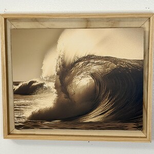 Puede incluir: Fotografía en tonos sepia de una gran ola oceánica, capturada en el momento de romper, enmarcada en un marco de madera clara. La cresta de la ola está iluminada, creando un contraste dramático con el agua más oscura debajo. La imagen evoca una sensación de poder y la belleza de la naturaleza.