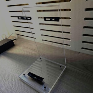 Pode incluir: Suporte de exibição em acrílico transparente com base retangular e painel vertical. O suporte possui hardware preto e foi projetado para segurar um item. O suporte está em uma superfície de madeira.