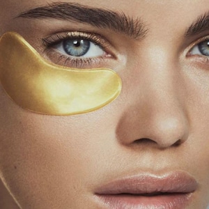 May include: Close-up of a person's face with blue eyes and a gold eye patch. The eye patch is a curved shape, covering the under-eye area. The skin has a smooth texture, and the eyebrows are dark and well-defined. The lips are a natural pink color.