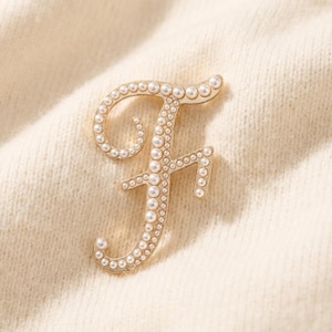 Puede incluir: Broche dorado con forma de letra "F", adornado con perlas de imitación. La broche se encuentra sobre una tela color crema, creando un contraste que resalta el intrincado diseño. Un elegante accesorio para un toque de sofisticación.