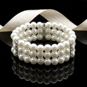 May include: A three-strand pearl bracelet with a silver clasp and rhinestone accents. The bracelet is made of round, white pearls and is displayed on a reflective black surface with a cream-colored ribbon in the background.