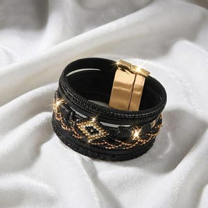 May include: Black cuff bracelet with gold-tone accents. The bracelet features multiple layers, including beaded and chain details. A gold-tone clasp secures the bracelet. The bracelet is displayed on a white, silky surface.
