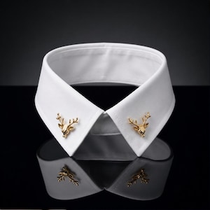 May include: A crisp white shirt collar featuring two gold-toned deer head pins attached to the points. The pins are shaped like stag heads with detailed antlers, providing a metallic accent to the plain white fabric of the structured collar.