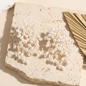 May include: A pair of elegant, gold-toned wire earrings, each adorned with clusters of white, oval-shaped pearls. The earrings have a floral design, resembling delicate branches. The earrings are displayed on a textured, light-colored surface.