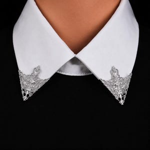 May include: A pair of silver-toned metal collar tips featuring an intricate, openwork filigree pattern. These decorative triangular accents are attached to the points of a crisp white shirt collar, contrasting against a solid black garment.