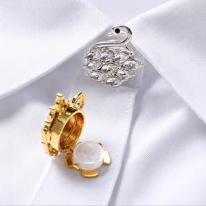 May include: A silver-toned swan-shaped brooch adorned with clear crystals and a gold-toned circular button cover with a white centre insert, both positioned on a white collared shirt fabric. The swan features a curved neck and crystal body.