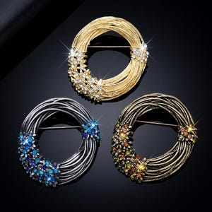 May include: Three decorative brooches in gold, silver, and bronze tones. Each brooch features a circular design made of layered metal strands, adorned with clusters of sparkling crystals in various colors. The brooches are set against a dark background.