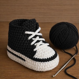 Może przedstawiać: Ręcznie robiony szydełkowy bucik dla dziecka w kolorze czarno-białym. But ma białe sznurowadła, biały nosek i białą podeszwę. Kłębek czarnej przędzy i szydełko leżą obok buta na drewnianej powierzchni.