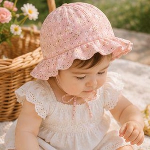 Puede incluir: Un sombrero de sol rosa claro con un delicado estampado floral blanco, una copa redondeada y un ala con volantes. El sombrero incluye cintas de tela a juego que se atan en un lazo bajo la barbilla, proporcionando un ajuste seguro en la cabeza.
