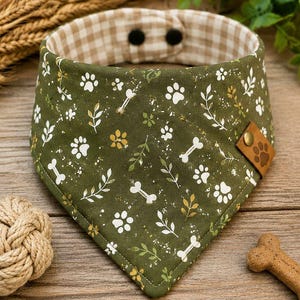 May include: A triangular pet bandana in olive green featuring a white pattern of paw prints, bones, and leafy sprigs. The interior is lined with a beige and white gingham check fabric and includes two black snap buttons for closure and a brown leather tag.