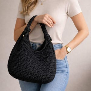 May include: A black, crocheted hobo bag with knotted handles and gold-colored hardware. The bag is held by a person wearing a cream-colored shirt and blue jeans. The bag's texture is visible, and the overall style is casual and handmade.